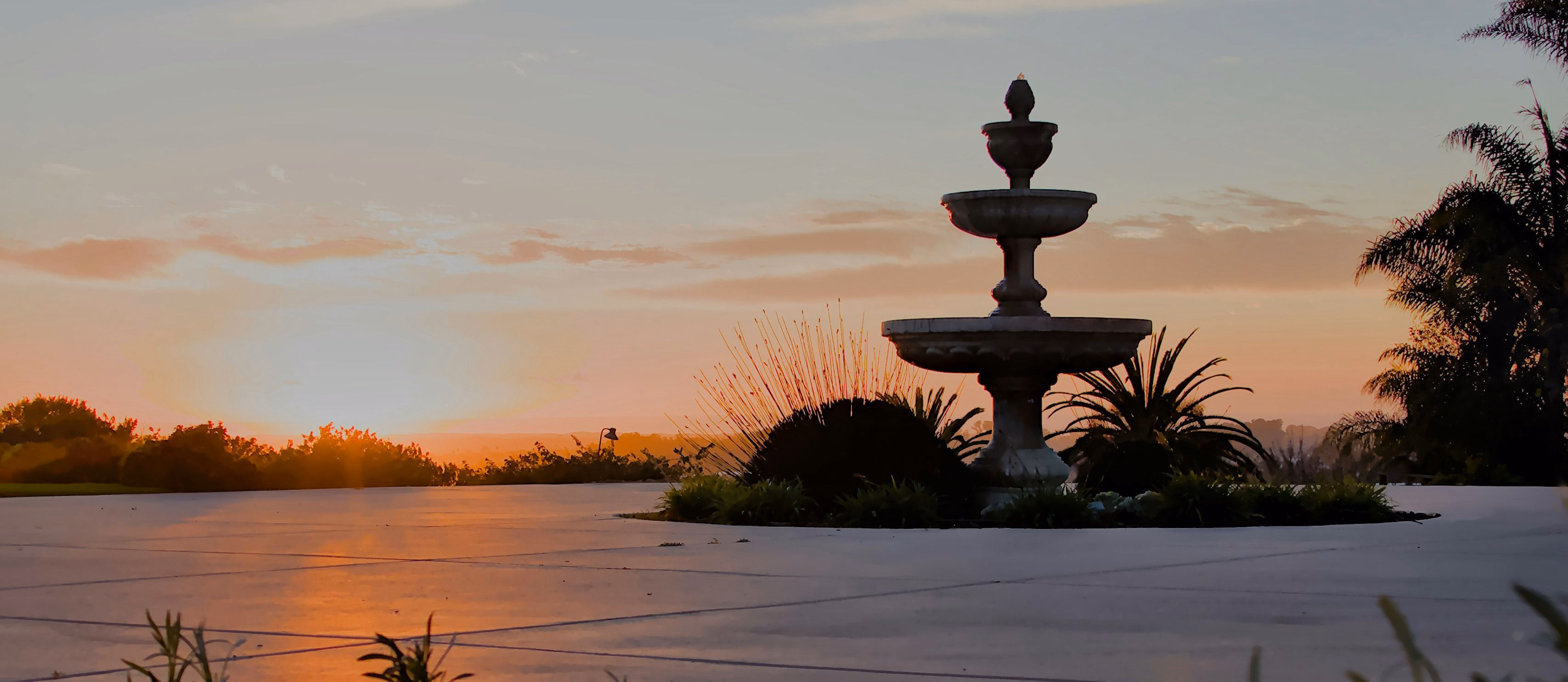 A tiered stone fountain on an open plaza at sunset, with palms and low hills on the horizon.